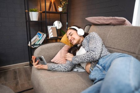 Foto de Feliz mujer joven relajada usando el teléfono móvil y los auriculares blancos mientras está acostado en el sofá. - Imagen libre de derechos