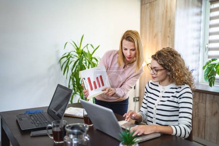 Photo for Two female colleagues are working from home, one is sitting at a desk using a laptop, while the other is standing next to her with a chart in hand, working together on a business report. - Royalty Free Image