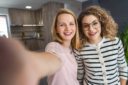 Zwei Frauen stehen vor einer Kamera, halten ein Handy in die Höhe und lächeln, als sie gemeinsam ein Selfie machen. 