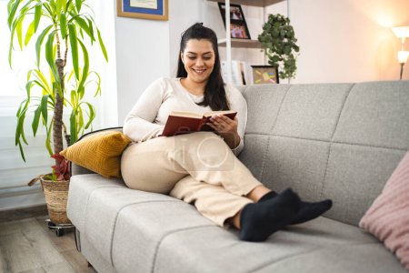 Foto de Mujer leyendo un libro mientras se relaja en un sofá en casa. - Imagen libre de derechos