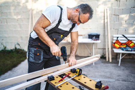 Téléchargez les photos : Mature mâle charpentier à l'aide d'une perceuse électrique sur une planche en bois à l'extérieur. - en image libre de droit