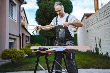 Téléchargez les photos : Mature mâle charpentier mesure planche en bois à l'extérieur. - en image libre de droit