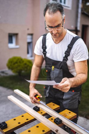 Photo for Mature male carpenter measuring wooden plank outdoors. - Royalty Free Image