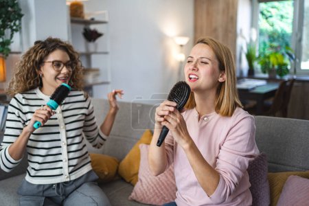 Téléchargez les photos : Deux amis sont assis sur le canapé et s'amusent à chanter dans des brosses à cheveux au lieu de microphones. - en image libre de droit