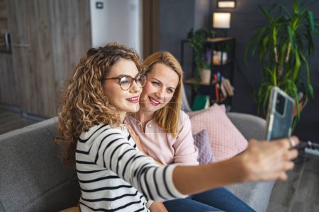 Deux femmes sont assises sur un canapé, tenant un smartphone à distance, souriant alors qu'elles prennent un selfie ensemble.