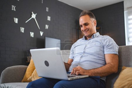 Téléchargez les photos : Moyen homme caucasien d'âge en utilisant un ordinateur portable sur le canapé. - en image libre de droit