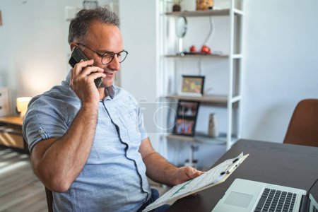 Photo for Middle aged Caucasian man talking on the phone while working on laptop at home. - Royalty Free Image