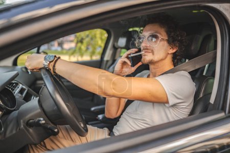 Téléchargez les photos : Homme aux cheveux bouclés et lunettes de soleil, conduit une voiture, fume une cigarette et utilise le téléphone. - en image libre de droit
