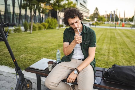 Foto de El hombre con el pelo rizado se sienta en un banco del parque, enciende un cigarro y descansa después de montar un scooter eléctrico - Imagen libre de derechos
