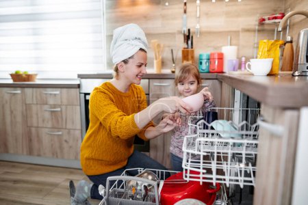 Foto de La niña ayuda a su mamá a cargar el lavavajillas en la cocina. Ella está muy orgullosa y feliz de ayudar a su madre en la cocina. - Imagen libre de derechos