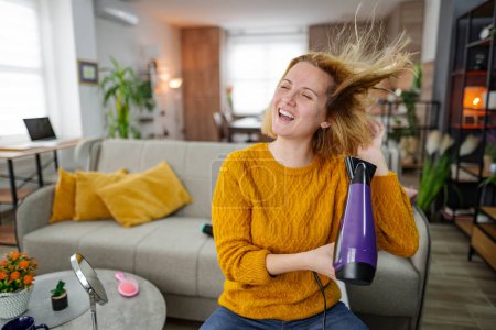 Photo for A woman is seated on a couch while holding a hair dryer in her hand. She appears to be in the middle of using the hair dryer, possibly grooming her hair or drying it after a shower. - Royalty Free Image