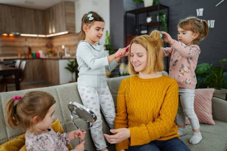 Photo for Three little girls are doing their moms hair on the couch in the living room, laughing and enjoying playing with their mom. - Royalty Free Image