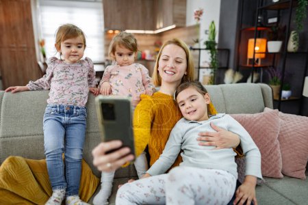 Téléchargez les photos : Mère et ses trois filles sont assises sur le canapé et prennent un selfie dans le salon à la maison. - en image libre de droit