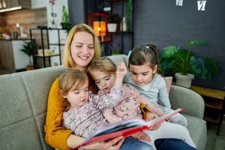 Téléchargez les photos : Mère et ses trois filles sont assises sur le canapé dans le salon, écoutant attentivement leur mère lire une histoire. - en image libre de droit