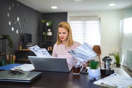 Téléchargez les photos : Femme blonde travaillant avec beaucoup de paperasse, assise à la table à la maison et très tendue - en image libre de droit
