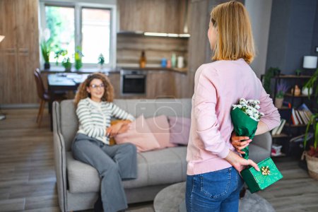 Foto de Una mujer rubia sonriente sorprende a su mejor amiga con un regalo y flores, de pie en medio de la sala de estar escondiendo el regalo y un ramo blanco detrás de su espalda. - Imagen libre de derechos