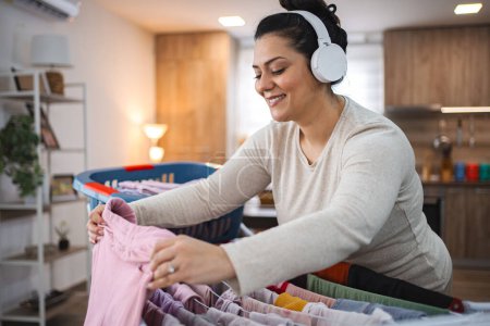 Téléchargez les photos : Femme écoutant de la musique tout en accrochant la lessive sur le séchoir à la maison. - en image libre de droit