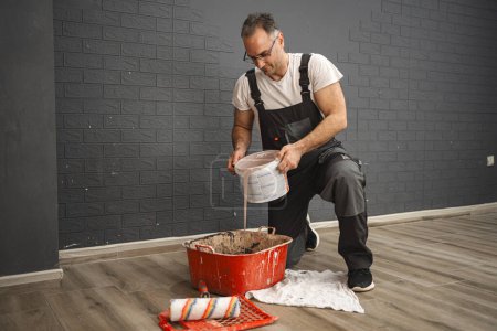 Photo for A kneeling man pours paint into a large plastic container. He wears work clothes and seems focused on the task at hand. - Royalty Free Image