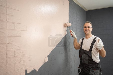 Foto de Un hombre está parado frente a una pared de ladrillo, pintándola diligentemente con un rodillo de pintura. Se centra en su tarea, extendiendo la pintura uniformemente sobre la superficie rugosa. - Imagen libre de derechos