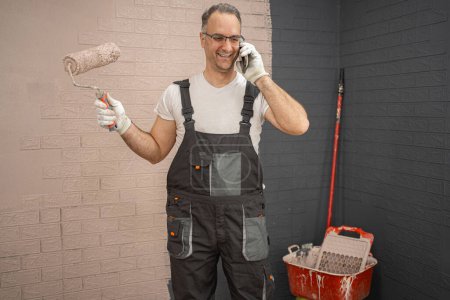 Man in overalls is standing and holding a roller in his hand. He is smiling and on the phone.