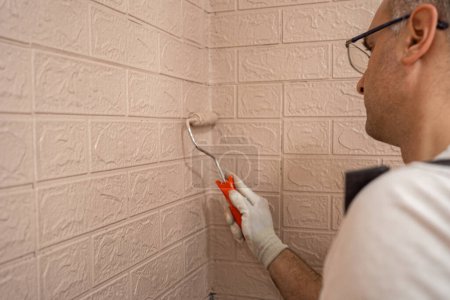 Un homme portant des gants blancs peint un mur de briques avec un petit rouleau de peinture. L'homme semble concentré sur la tâche, couvrant soigneusement la surface avec de la peinture.