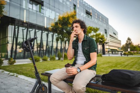 Téléchargez les photos : Homme aux cheveux bouclés assis sur un banc de parc, fume un cigare et se repose après avoir roulé un scooter électrique. - en image libre de droit