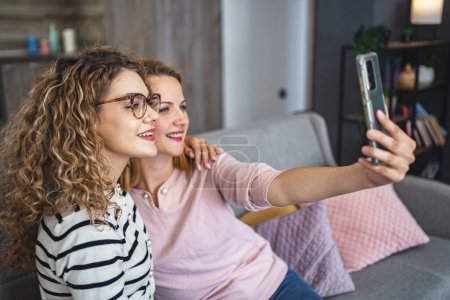 Deux femmes sont assises sur un canapé, tenant un smartphone à distance pour prendre un selfie ensemble. Ils sourient tout en capturant le moment sur l'écran.