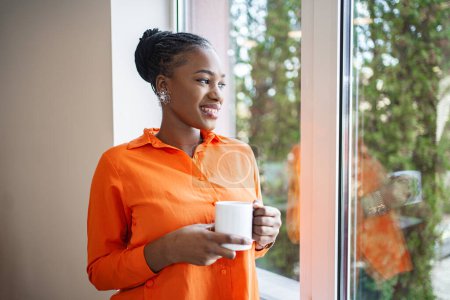 Téléchargez les photos : Une femme portant une chemise orange tient une tasse dans sa main. Elle se tient dans un endroit inconnu, regardant vers la caméra. - en image libre de droit