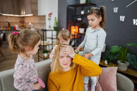 Téléchargez les photos : Trois petites filles font leurs cheveux de mamans sur le canapé dans le salon, riant et appréciant jouer avec leur mère. - en image libre de droit