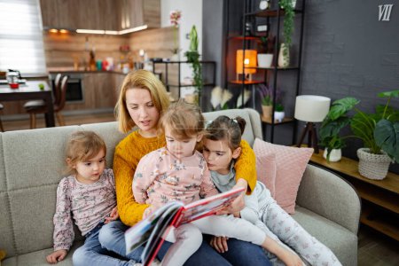Téléchargez les photos : Une mère et ses trois filles sont assises sur le canapé dans le salon, écoutant attentivement leur mère lire une histoire. - en image libre de droit