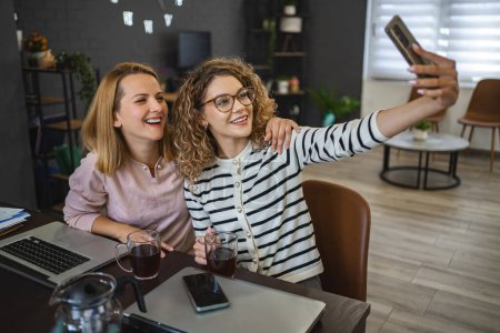 Photo for Two female colleagues are sitting at the table with a cup of coffee and taking a selfie. - Royalty Free Image