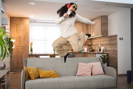 Téléchargez les photos : Femme avec casque sans fil sautant sur le canapé et chantant à la maison. - en image libre de droit