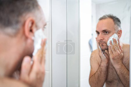 Foto de Un hombre se aplica espuma de afeitar en la cara en el baño. - Imagen libre de derechos