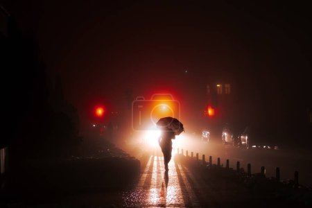 Ein Mann mit Regenschirm geht durch die nächtliche neblige Stadt. Die Strahlen von Autoscheinwerfern und Ampeln erhellen die Silhouette einer Person. Weicher Fokus. Hochwertiges Foto