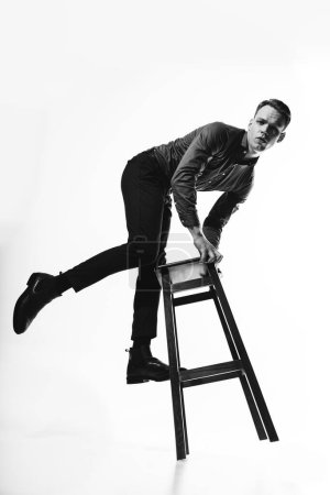 Foto de Un elegante retrato en blanco y negro de un hombre con una camisa posando sobre un ciclama blanco con una silla, evocando una sensación de sofisticación y refinamiento. Foto de alta calidad - Imagen libre de derechos