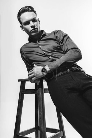 Foto de Un elegante retrato en blanco y negro de un hombre con una camisa posando sobre un ciclama blanco con una silla, evocando una sensación de sofisticación y refinamiento. Foto de alta calidad - Imagen libre de derechos