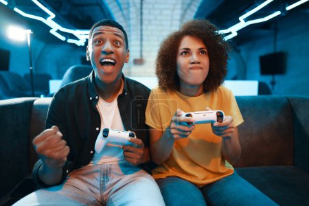 Téléchargez les photos : Un homme et une femme sont assis sur un canapé à jouer à un jeu vidéo, profiter du temps libre et s'amuser dans un événement à thème bleu électrique - en image libre de droit