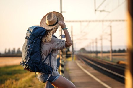 Train traveler with backpack and hat is waiting at railroad station platform. Woman traveling on vacation alone