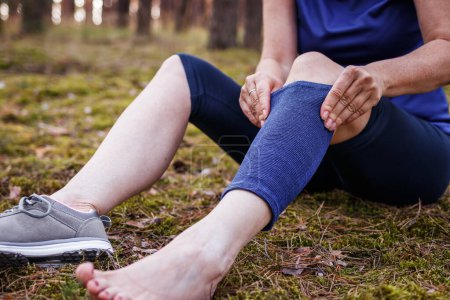 Foto de Caminante poniendo vendaje ortopédico elástico durante el senderismo. Mujer sintiendo dolor de articulación y rodilla después de una lesión. Primeros auxilios de ligamento desgarrado o esguince de rodilla fijación al aire libre - Imagen libre de derechos