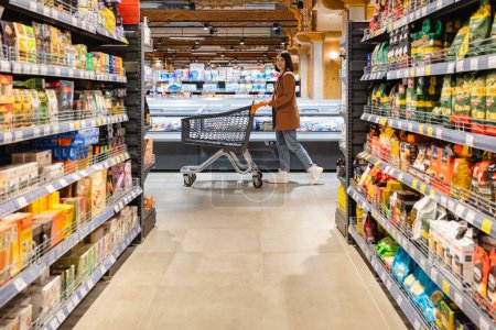 une femme avec un chariot marche entre des rangées d'étagères dans une épicerie