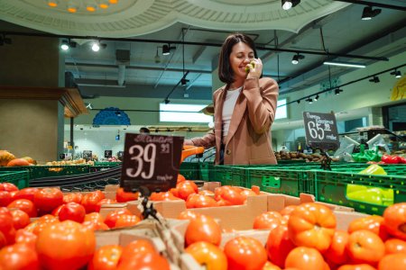 Foto de Retrato de una dama milenaria comprando comida, caminando en un supermercado con un carro. Una cliente hembra huele tomates mientras toma productos de estantes en una tienda - Imagen libre de derechos