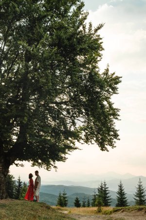 Photo for A couple stands and looks at each other on a bench by a big old beech tree with a view of the carpathians mountains - Royalty Free Image