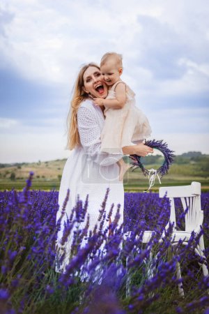 Eine glückliche Mutter umarmt ihre Tochter und lächelt, während sie neben einem weißen Stuhl in einem Lavendelfeld steht