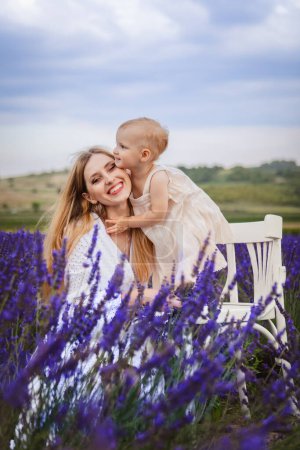 In a lavender field, a mother hugs her daughter as she stands on a white chair.