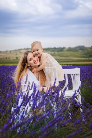Auf einem weißen Stuhl in einem Lavendelfeld stehend, umarmt eine glückliche Mutter ihre Tochter auf einem weißen Stuhl