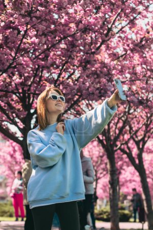 Téléchargez les photos : Femme heureuse prenant selfie sur le téléphone en face de l'espace de copie sakura floraison - en image libre de droit