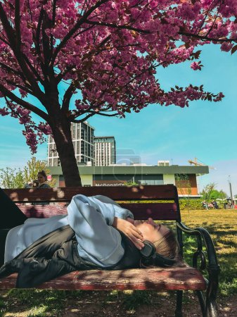 Téléchargez les photos : Femme allongée sur le banc écoutant de la musique sous un sakura en fleurs - en image libre de droit