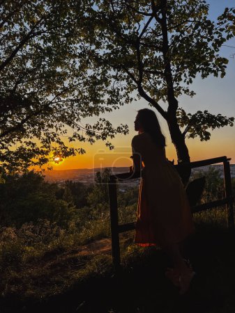 Téléchargez les photos : Coucher de soleil vista : femme par clôture, regardant le paysage urbain - en image libre de droit