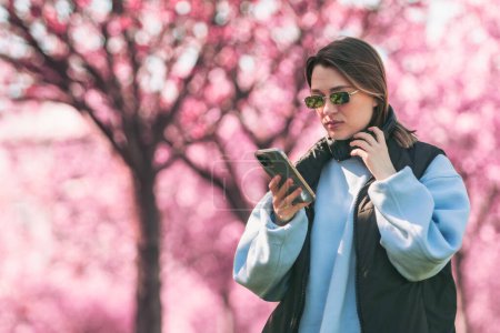 Téléchargez les photos : Femme amateur de musique vérifier des chansons sur son téléphone copier l'espace sakura en fleurs sur fond - en image libre de droit