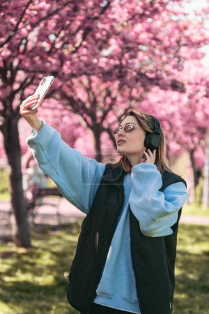 Téléchargez les photos : Femme heureuse écouter de la musique dans le casque à l'extérieur en prenant selfie avec sakura sur l'espace de copie de fond - en image libre de droit